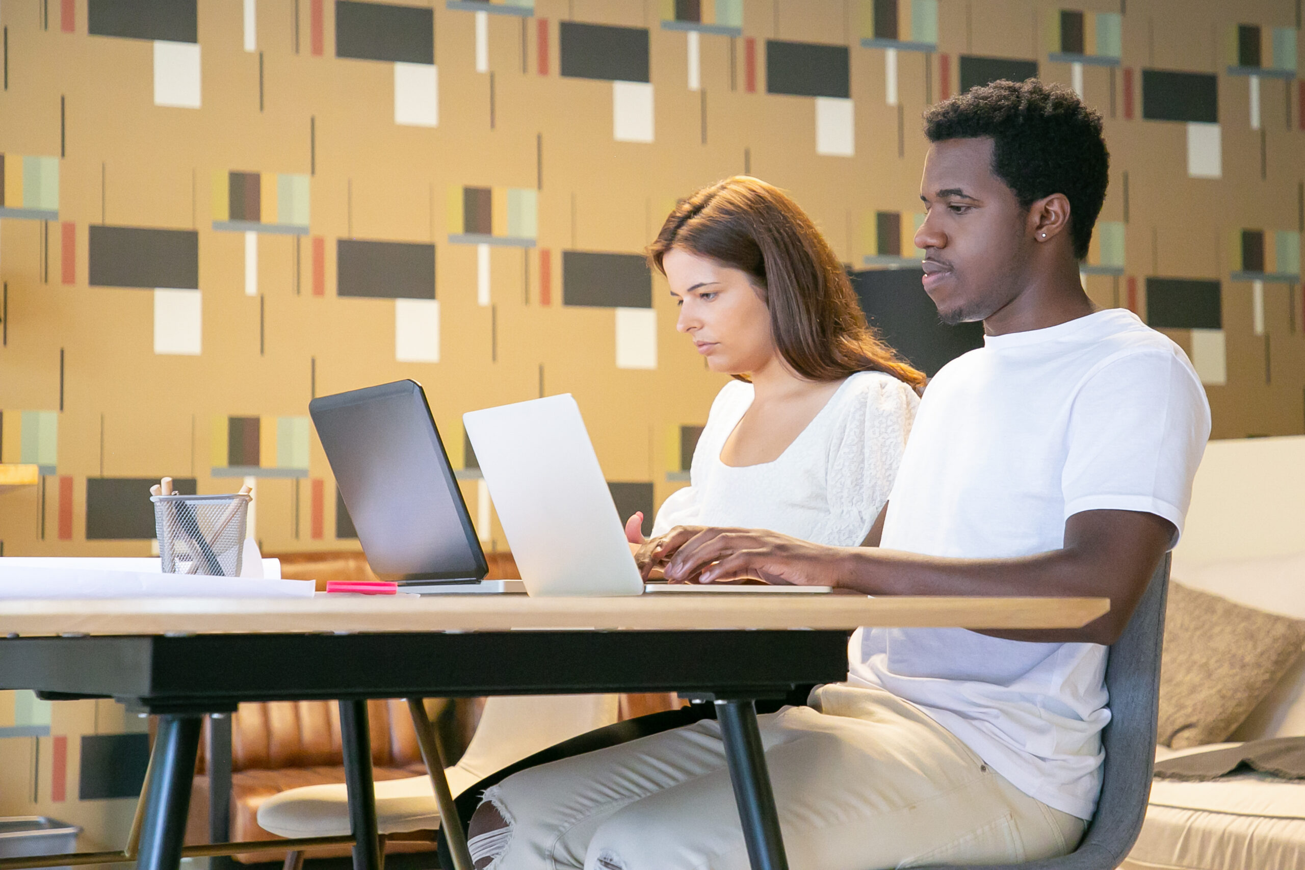 Couple of creative professionals sitting together at table with blueprints and working on project. Two colleagues typing on laptops. Coworking office concept
